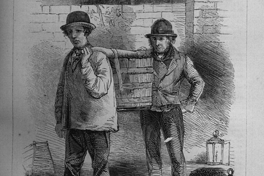 A historical black-and-white engraving depicting two men in worn clothing and bowler hats carrying a large wooden basket suspended on a pole over one man's shoulder. The second man stands with one hand on his hip, giving the impression of a working-class profession. A lantern and another basket rest on the ground, suggesting they might be street vendors or laborers in an urban setting.