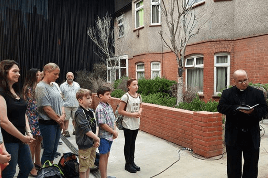 A group of people, including adults and children, stand solemnly in an outdoor courtyard while a priest in black robes reads from a book. The setting features a brick building with windows and leafless trees, with some greenery in the background. The expressions of the attendees convey seriousness and reverence, suggesting a memorial or blessing ceremony.