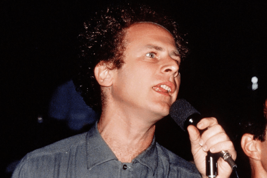 A male singer passionately performs, holding a microphone close to his mouth while singing with an intense expression. He wears a casual button-up shirt, and his curly hair frames his face as he leans into the performance. The dark background with blurred figures suggests a live concert or intimate venue setting.