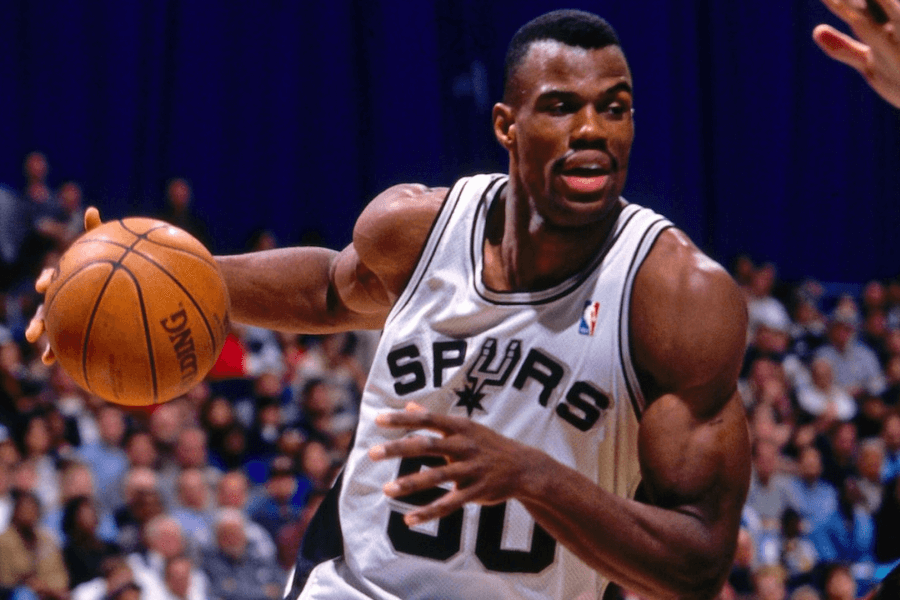 A basketball player wearing a San Antonio Spurs jersey with the number 50 is in action, dribbling the ball while looking ahead. His muscular build and determined expression highlight his athleticism as he moves across the court. The background shows a blurred crowd of spectators, emphasizing the intensity of the game.