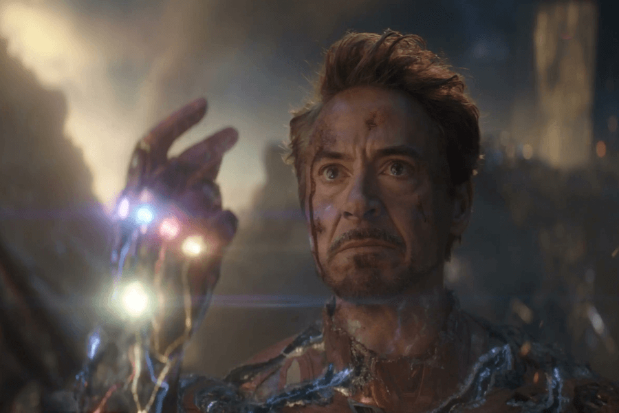 A battle-worn man with a determined expression and a bruised face raises his hand, which is glowing with the power of six embedded, multicolored stones. His armor is damaged, revealing parts of his body as energy surges through him. The dramatic lighting and blurred battlefield background emphasize the intensity of this pivotal moment.