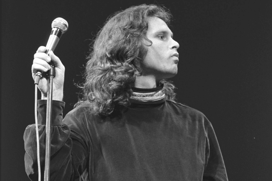 A black and white photograph of a male rock singer with wavy, shoulder-length hair standing on stage. He holds a microphone in one hand while gazing off to the side with an intense and contemplative expression. His dark, loose-fitting velvet shirt and high collar add to his enigmatic stage presence.