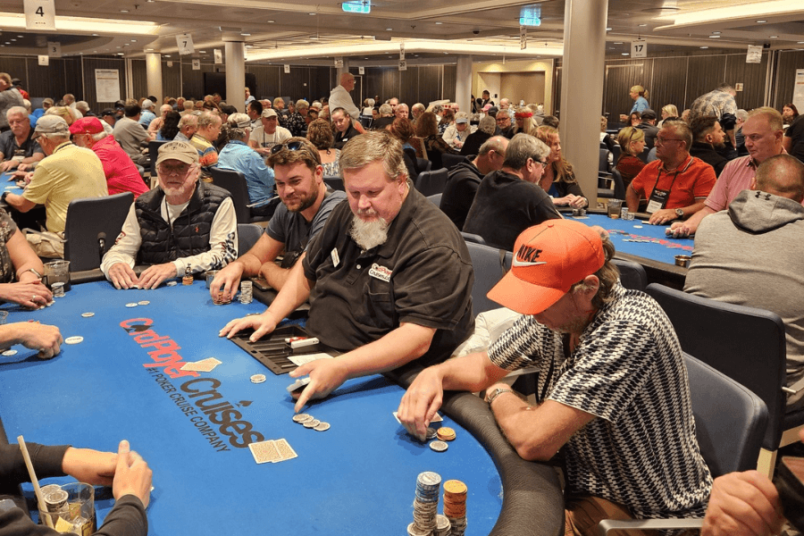 A lively poker tournament aboard a cruise ship, with players seated around a blue felt table labeled "Card Player Cruises." A dealer distributes chips while participants, including a man in a bright orange Nike cap, focus on their hands. The room is filled with additional poker tables, and the background shows a crowded, engaged atmosphere of players enjoying the competition.