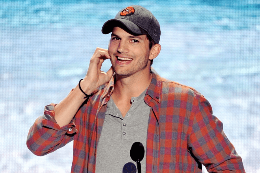 A man in a casual outfit stands on stage, smiling warmly while scratching the side of his face. He wears a gray baseball cap, an orange and gray plaid shirt over a buttoned-up gray T-shirt, and a black wristband. The background features a large screen displaying an ocean-like scene, giving the setting a relaxed and fun atmosphere.