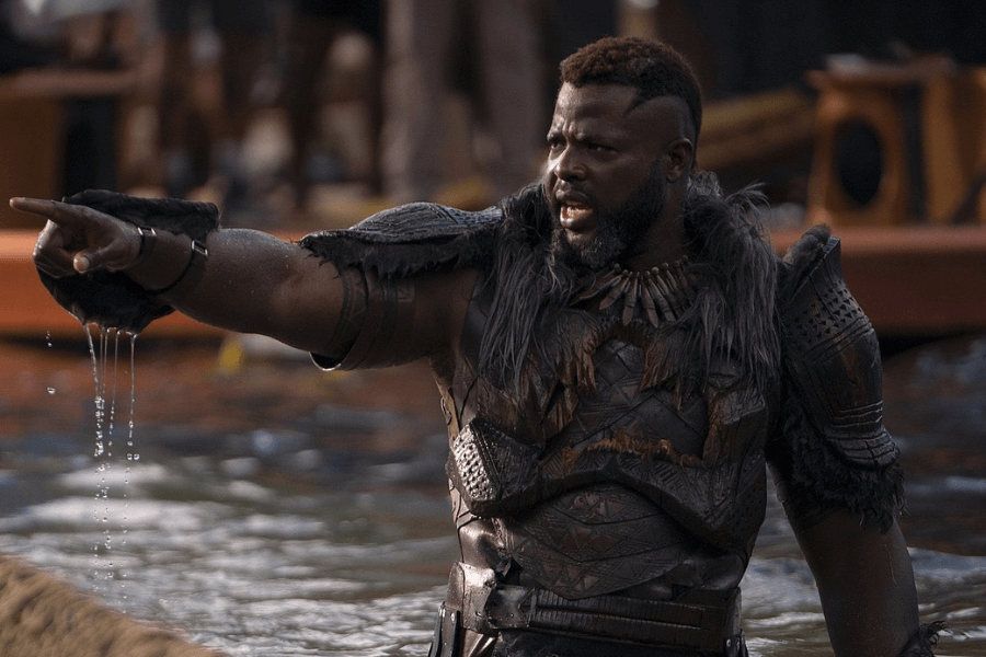 A powerful warrior stands in the water, pointing forcefully with a determined and commanding expression. He wears fur-trimmed, intricately designed armor with tribal elements, and water drips from his outstretched arm. Behind him, wooden boats and a gathering of people hint at a significant ritual or challenge.