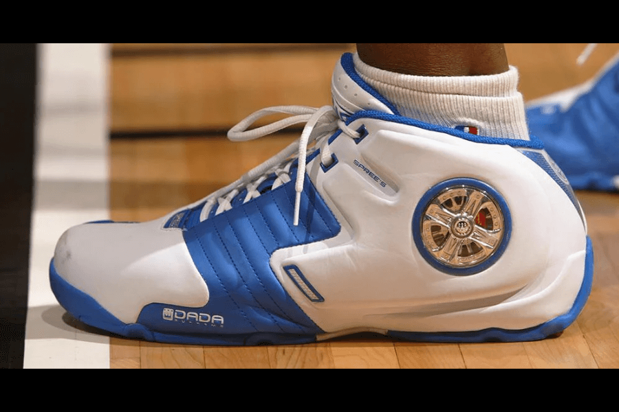 A close-up of the iconic Dada Supreme Spinners basketball sneaker in white and blue, featuring a built-in chrome spinning rim detail on the heel. The shoe has a sleek, padded design with quilted side panels and bold "Dada Supreme" branding near the sole. Known for its flashy, over-the-top aesthetic, this sneaker became a legendary piece of early 2000s basketball and hip-hop fashion.