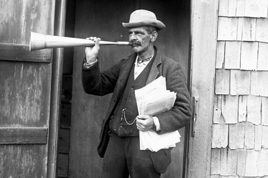 A historical black-and-white photograph of a town crier or news announcer standing outside a wooden building. He wears a worn suit, vest, and a bowler hat, holding a stack of newspapers in one hand while using a large metal horn to amplify his voice. His serious expression and posture suggest he is delivering an important announcement to the community.