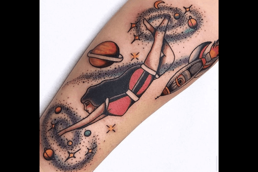 A colorful tattoo featuring a retro-inspired female figure in a swimsuit floating through space, surrounded by swirling galaxies, planets, and stars. A rocket ship is depicted in motion on the right, adding to the whimsical, cosmic theme. The design combines vibrant hues and playful details to create a dreamlike, vintage aesthetic.