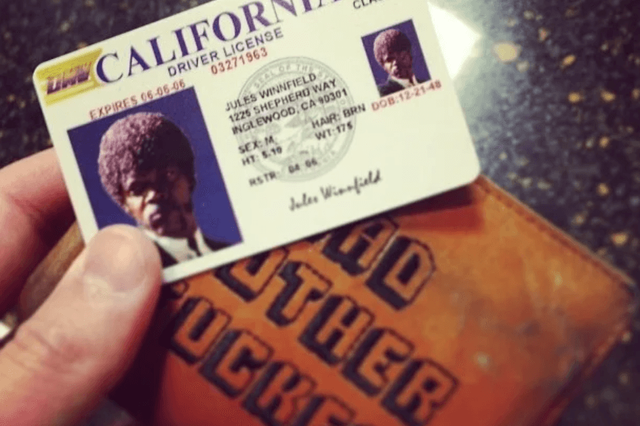 A hand holds up a novelty California driver's license featuring the character Jules Winnfield from Pulp Fiction, with his signature afro hairstyle and intense expression. The license is placed above a brown leather wallet embossed with bold black text. The humorous and cinematic reference adds to the nostalgic and pop culture appeal of the image.