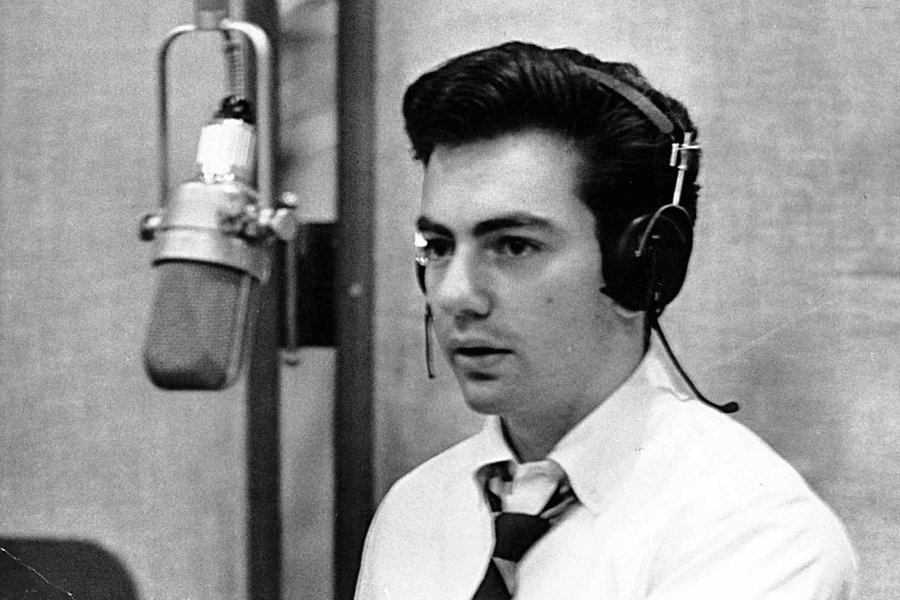 A black and white photograph of a young male singer in a recording studio, wearing headphones and a slightly loosened tie. He looks focused as he sits in front of a vintage microphone, seemingly preparing for a take. The studio's textured walls and classic equipment create a timeless atmosphere.