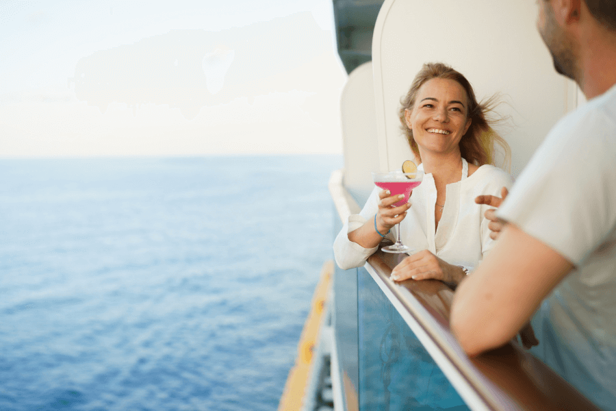 A smiling woman leans on the railing of a cruise ship balcony, holding a pink cocktail garnished with a lime slice. She is engaged in conversation with a man, whose back is partially visible in the foreground. The background features the open ocean stretching to the horizon under a clear sky.