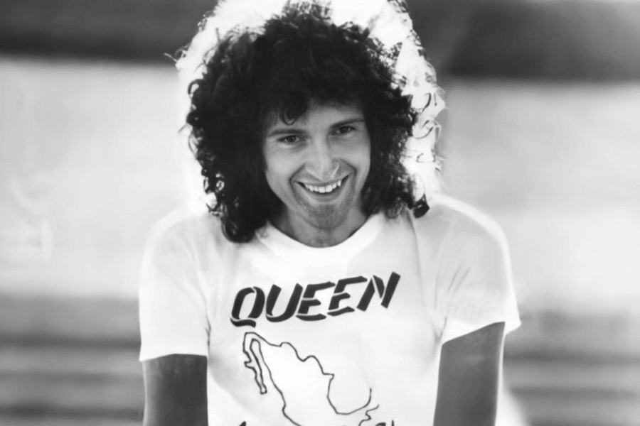 A black-and-white photograph of a man with curly hair smiling warmly at the camera. He wears a white T-shirt with "QUEEN" printed in bold letters and a graphic design underneath. The soft lighting and blurred background create a nostalgic and vintage rock-and-roll feel.