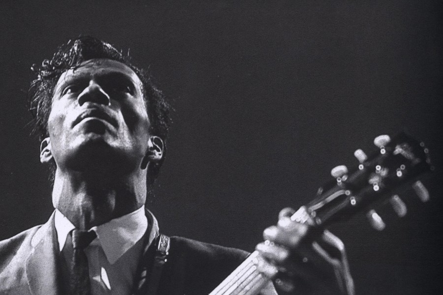 A black and white photograph captures a male guitarist in a dramatic low-angle shot, looking upward with an intense expression. He wears a suit and tie, gripping his guitar as stage lighting casts deep shadows on his face. The image exudes a powerful and iconic rock and roll presence.