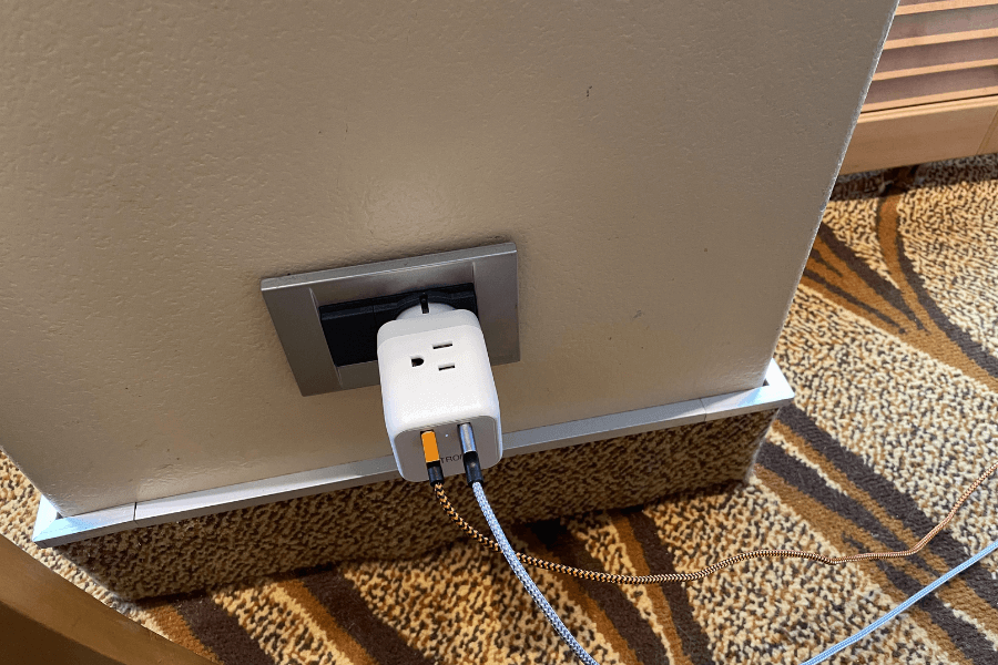 A cruise ship power outlet with a white multi-port adapter plugged in, charging multiple devices using braided charging cables. The outlet is embedded in a beige wall with a metallic frame, and the patterned carpet below adds to the cozy interior setting. The setup highlights a common cruise travel necessity for charging multiple electronics.