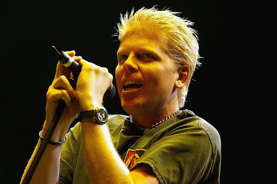 A blonde-haired man passionately sings into a microphone during a live concert. He wears a dark T-shirt, a wristwatch, and a chain necklace, gripping the mic stand with intensity. The dramatic stage lighting casts a warm glow on his face, emphasizing the energy of the performance.