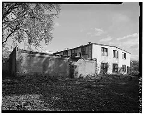 Black and white photo of an old, two-story building with cracked walls and overgrown surroundings. A large tree stands to the left, partially shading the structure under a mostly clear sky