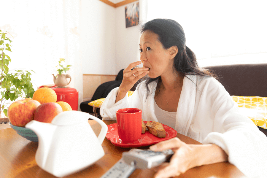 A woman in a white robe sits at a wooden table, eating breakfast while holding a remote control. A red cup, a plate with toast, and a bowl of fruit are on the table, with a cozy and well-lit living space in the background.