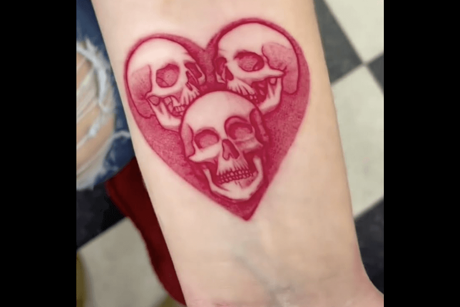 A striking tattoo of three detailed skulls enclosed within a bold, red heart shape. The skulls are shaded in monochromatic tones of red, blending seamlessly into the heart’s textured design, creating a cohesive and dramatic visual effect. The composition conveys a balance between macabre and romantic symbolism.