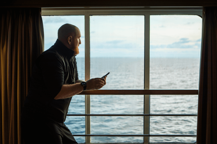 A man with a beard and wearing a black sweater leans on the railing of his cruise ship cabin's balcony, gazing out at the vast ocean. He holds a smartphone in one hand, seemingly lost in thought, while the soft natural light from the window highlights the calm, open sea stretching to the horizon. The moment captures a peaceful and reflective mood.