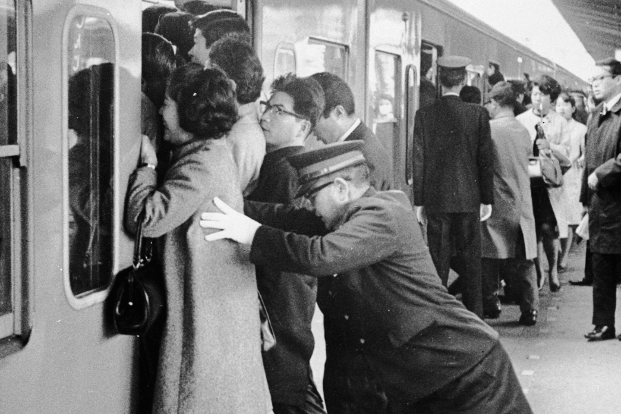 A black-and-white historical photograph capturing a crowded train station, where a uniformed railway worker is seen pushing passengers into an already packed train. The commuters, dressed in mid-20th-century attire, struggle to fit inside as other station staff assist. The image reflects the extreme rush-hour congestion typical of urban transit systems, likely in Japan.
