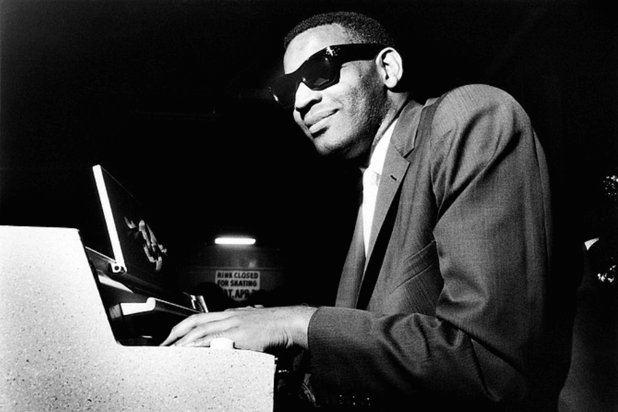 A black and white photograph of a pianist wearing dark sunglasses and a suit, playing an electric keyboard with a confident smile. The lighting casts a dramatic contrast, highlighting his expressive face and hands on the keys. A sign in the background reads "RINK CLOSED FOR SKATING," adding a subtle atmospheric detail.