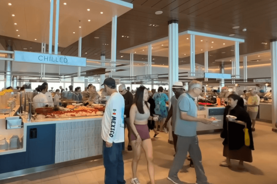 A spacious cruise ship buffet with passengers selecting food from various stations. The "Chilled" section is visible in the foreground, featuring seafood and fresh ingredients, while other food stations, including "Indian" and "Latin," are seen in the background. Passengers in casual attire walk around with plates, engaging in conversations and enjoying the dining experience.