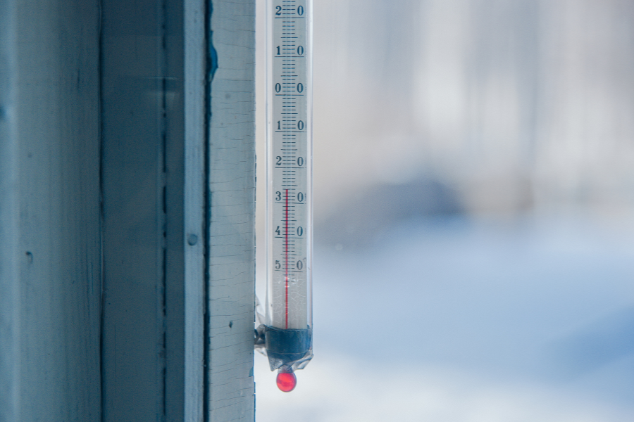 A glass thermometer mounted outside a window shows a temperature well below freezing, with snow-covered scenery in the blurred background. The red liquid inside the thermometer is near the bottom, indicating extremely cold weather.