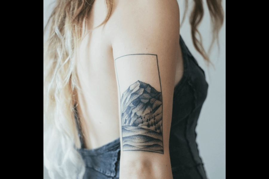 A minimalistic tattoo featuring a geometric rectangle frame containing a mountain landscape. The scene is composed of sharp-edged peaks, rolling hills, and a dot-work shading technique.