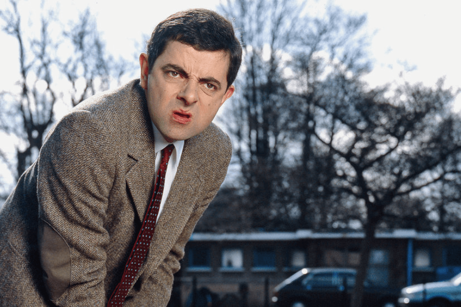 A man with dark hair and exaggerated facial features squints and makes a puzzled expression while leaning forward slightly. He wears a brown tweed jacket, a white shirt, and a red tie, standing outdoors with leafless trees and parked cars in the background. His comical expression suggests confusion or curiosity.