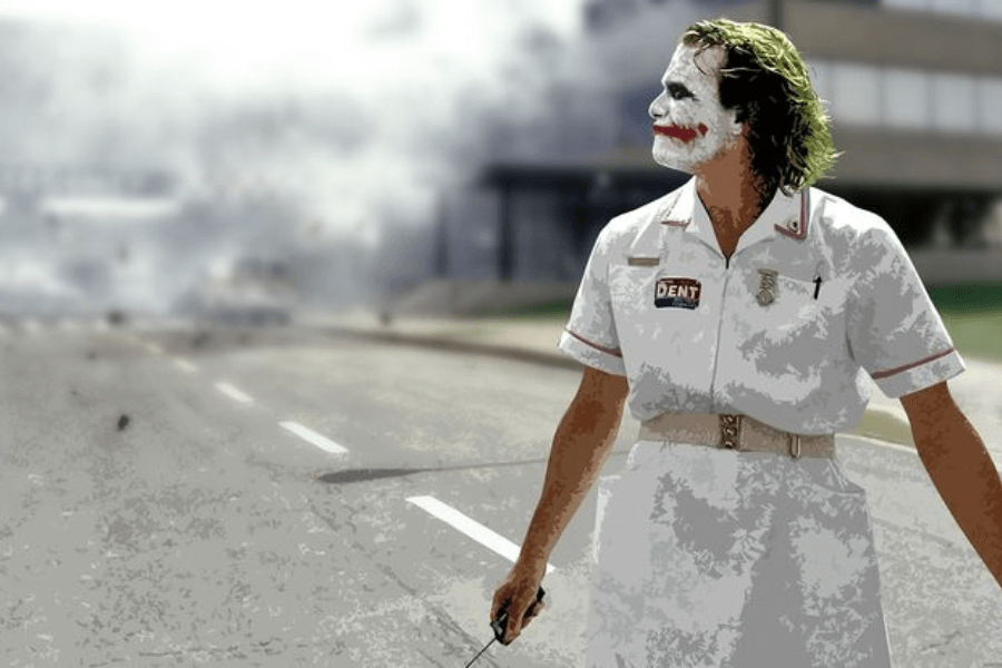 A man with white face paint, red smeared lips, and green disheveled hair wears a white nurse's uniform with a "Dent" name tag. He stands in the middle of a desolate road holding a detonator, looking off to the side while an explosion engulfs the background in smoke and debris. The dramatic lighting and stylized effect give the scene an intense, cinematic feel.