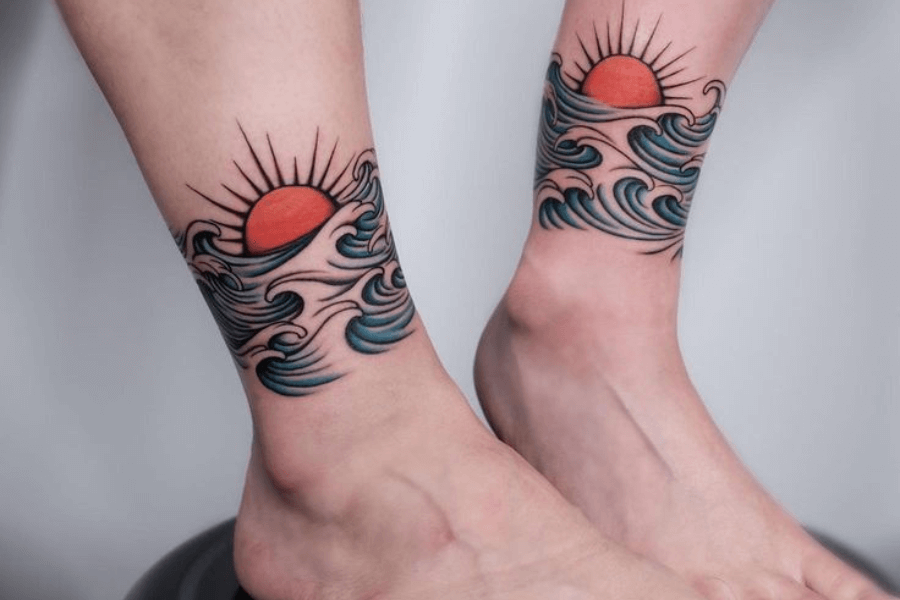 A vibrant tattoo spanning both ankles, featuring a red sun rising or setting above stylized, rolling ocean waves. The waves are detailed with bold black outlines and shades of blue, while the sun radiates thin black rays, creating a balanced and symmetrical design. This artistic piece evokes a sense of serenity and connection with nature.