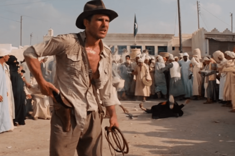 A rugged adventurer in a dusty fedora and unbuttoned shirt stands in a bustling marketplace, holstering his pistol while gripping a whip. A fallen swordsman lies in the dirt behind him as a shocked crowd in traditional Middle Eastern attire watches. The scene is filled with dust and movement, capturing an intense moment of quick action.