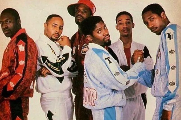 A classic 80s hip-hop group photo featuring six men dressed in colorful Troop tracksuits. The group members wear coordinated red, white, and blue athletic sets adorned with the Troop logo and crown motifs, showcasing the bold streetwear style of the era. Two men in the foreground shake hands in a display of unity, while others pose with serious expressions, exuding confidence and swagger. The image embodies the golden age of hip-hop fashion and the dominance of Troop as a streetwear staple.