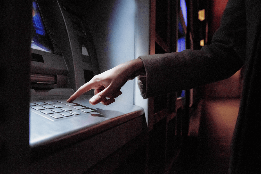 A close-up of a person using an ATM, with their hand pressing a button on the keypad. The dim lighting and shadows create a slightly moody atmosphere, while the screen of the ATM emits a faint glow. The person is wearing a long-sleeved coat, suggesting a cool or formal setting.

