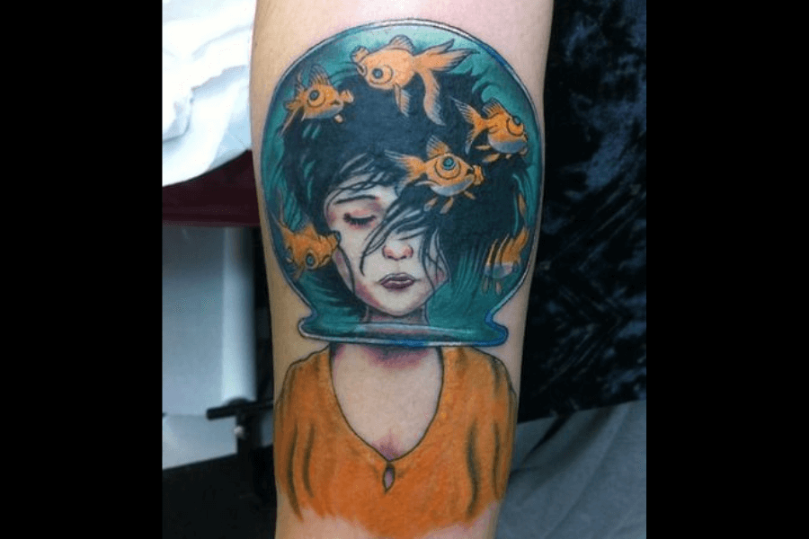 A vibrant tattoo of a woman with her head encased in a fishbowl, surrounded by goldfish swimming through her black hair. Her eyes are closed, and her expression is serene, wearing a warm orange blouse that contrasts with the cool tones of the water in the bowl. The design blends surrealism and vivid color to create a dreamlike and introspective atmosphere.