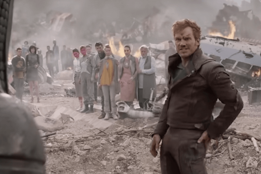 A battle-worn man in a dark leather jacket stands amidst the ruins of a destroyed city, his face bruised and tense. Behind him, a group of solemn-looking people, some with unusual skin colors, watch in concern. Flames and wreckage from a crashed spaceship burn in the background, adding to the chaotic scene.