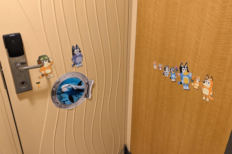 A decorated cruise cabin door featuring magnetic stickers of characters from the animated show Bluey and a faux porthole magnet with an image of an orca. The wavy-textured beige door has a keycard lock and a handle, while the adjacent wooden panel also has additional Bluey character magnets arranged in a line.