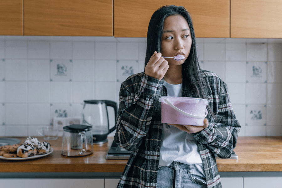 A young woman in a plaid shirt and jeans stands in a kitchen, eating directly from a large tub of yogurt with a spoon. She gazes off to the side with a thoughtful expression, while a plate of pastries and a coffee maker sit on the counter behind her.