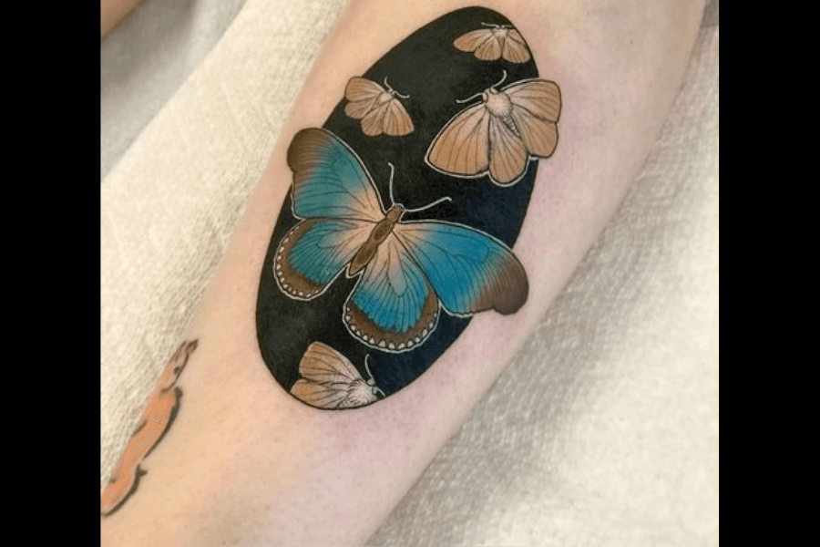 A tattoo featuring a vibrant blue butterfly as the centerpiece, surrounded by smaller beige moths, set against a solid black oval background. The intricate details of the wings highlight their texture and colors, creating a striking contrast with the dark backdrop. The design conveys a harmonious balance between beauty and simplicity.