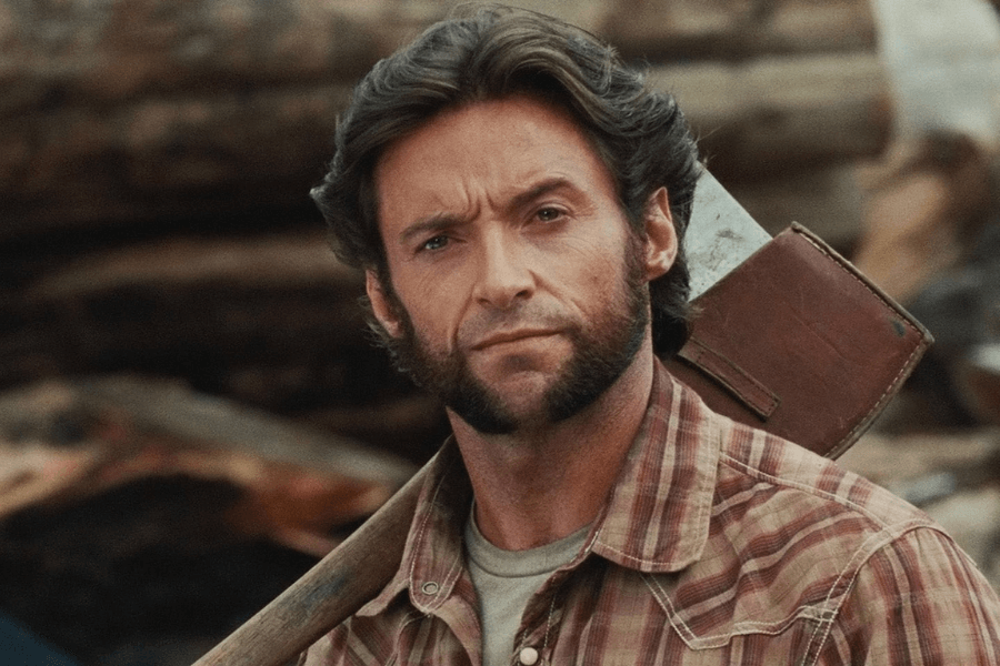 A rugged man with thick sideburns and tousled dark hair stands in a lumberyard, holding an axe over his shoulder. He wears a plaid flannel shirt and has an intense, serious expression. The background consists of stacked logs, emphasizing the outdoorsy, hardworking setting.