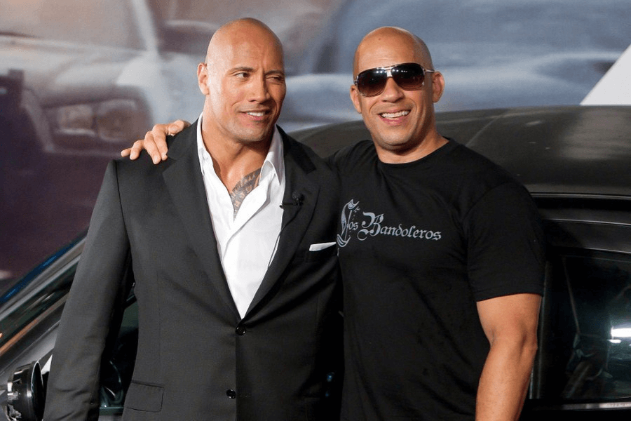 Two muscular, bald men pose together at a promotional event, smiling and looking confident. One is dressed in a black suit with a white shirt, while the other wears sunglasses and a black T-shirt with "Los Bandoleros" written on it. They stand in front of a sleek black car, with a blurred action-packed background.