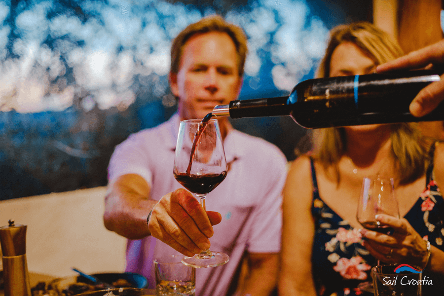 A man in a light pink polo shirt extends his hand to receive a glass of red wine as it is poured from a bottle. A woman in a floral dress sits beside him, holding her own wine glass. The setting is an intimate dining experience with a warm ambiance, and the logo "Sail Croatia" is visible in the bottom right corner, suggesting a wine-tasting event on a sailing trip.