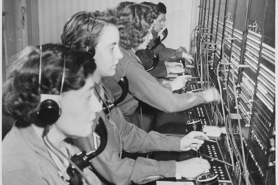 A black-and-white historical photograph of a row of female telephone operators working at a large switchboard. They are dressed in uniforms, wearing headsets with mouthpieces, and are seen efficiently plugging and unplugging cables to connect calls. The image reflects the mid-20th-century era, highlighting the vital role of women in communication industries.