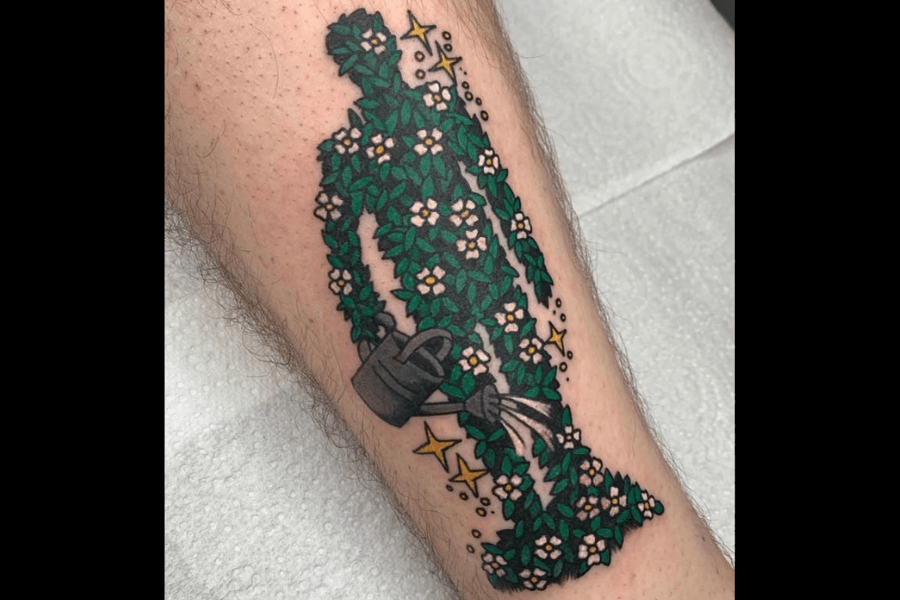 A tattoo depicting a silhouette of a human figure made entirely of green leaves and white flowers, with a black watering can at the base. Yellow stars and small decorative dots surround the design, adding a whimsical touch. The piece symbolizes growth, nature, and nurturing, blending bold outlines with intricate floral details.