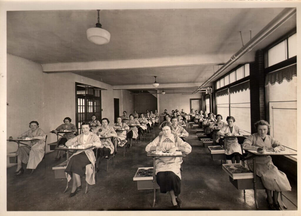 A sepia-toned photograph of a large group of women seated in neat rows at individual desks in a spacious factory or workshop. They are wearing smocks and appear to be engaged in precision work, possibly painting watch dials. The setting suggests an early-to-mid 20th-century industrial workplace, reminiscent of the 'Radium Girls' who worked with luminous radium paint.