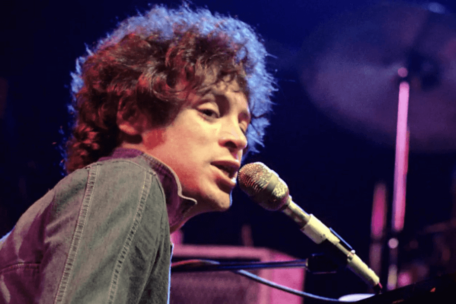 A curly-haired musician leans towards a gold-colored microphone while performing on stage. He is wearing a denim jacket, and the stage lighting casts a warm glow on his face. A drum set and other stage equipment are visible in the softly blurred background, adding to the live concert ambiance.