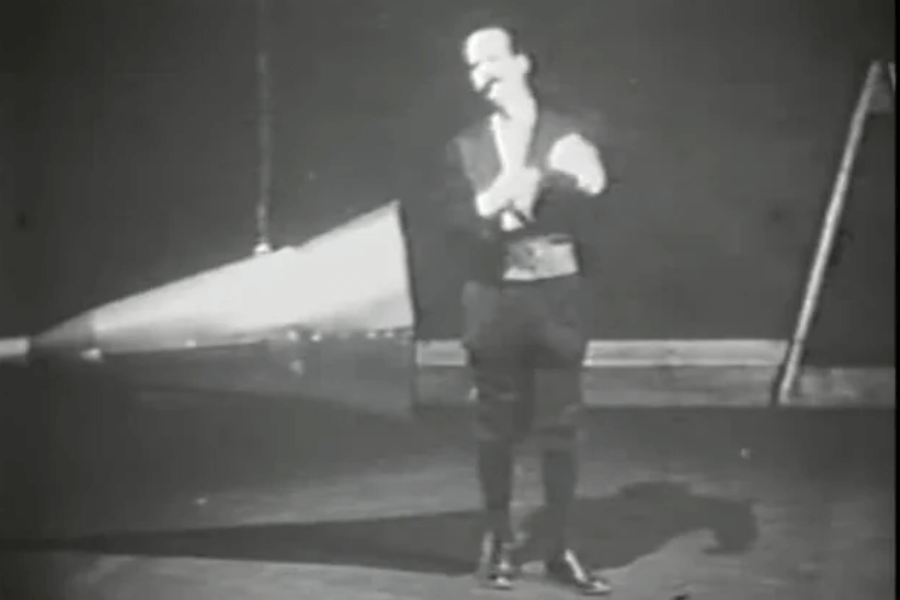  A grainy black-and-white image of a man in formal attire standing on a stage with a massive megaphone-like device behind him. The man, dressed in a suit with a vest, appears to be addressing an audience or preparing for a performance. The setting suggests an early 20th-century experiment or theatrical demonstration.