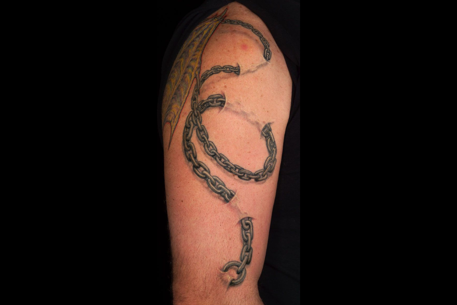 A tattoo featuring a realistic depiction of a broken chain wrapping around the upper arm. The chain links are shaded to create a three-dimensional effect, with jagged breaks symbolizing freedom or strength. The design is bold and impactful, blending seamlessly with the arm's natural contours.