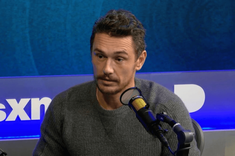 A man with short, wavy brown hair and a trimmed beard sits in a radio studio, speaking into a professional microphone. He wears a gray knit sweater and has a serious yet engaged expression. The blue-lit background features the "SXM" logo, indicating a SiriusXM interview setting.