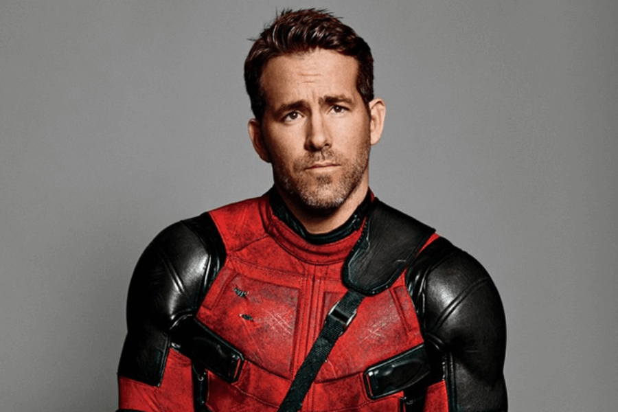 A man in a red and black superhero suit stands against a plain gray backdrop, giving a slightly smirking yet thoughtful expression. His suit is made of textured, battle-worn material with black shoulder armor and a strap crossing his chest.